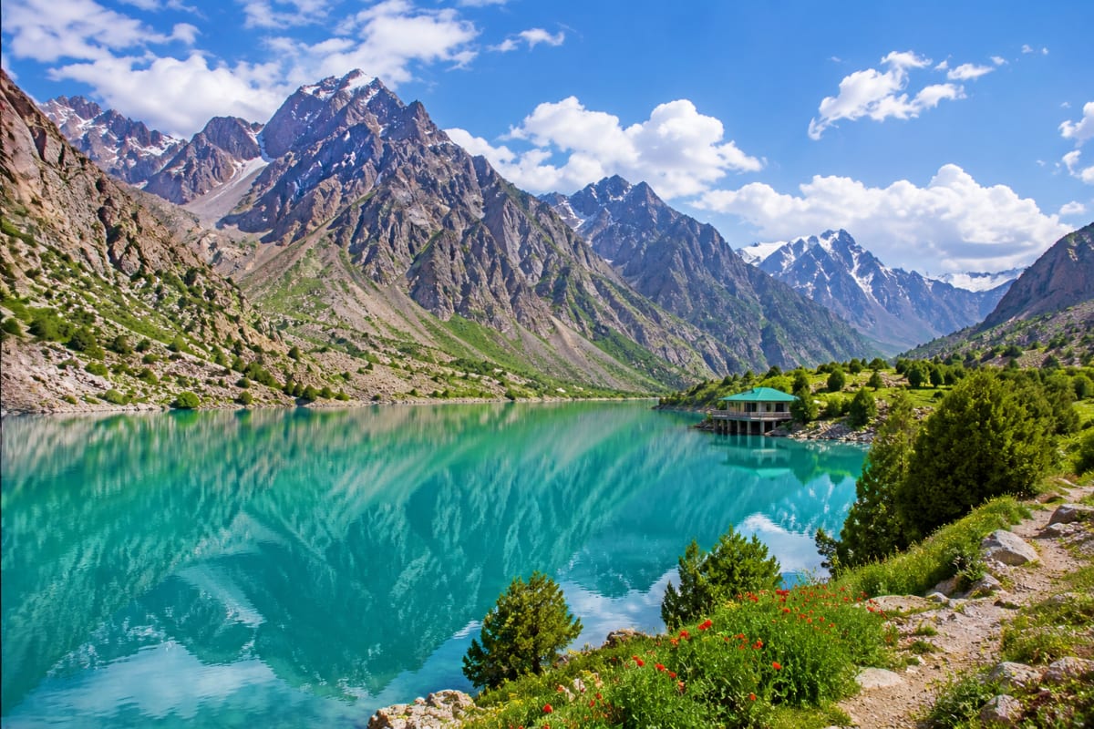 Tajikistan Visa Turquoise alpine lake surrounded by jagged mountain peaks and green vegetation in Tajikistan with traditional structure on sh
