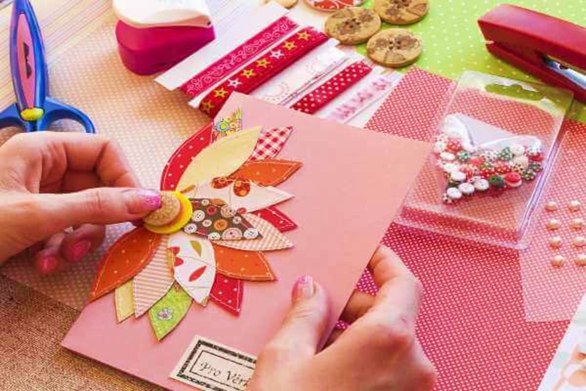 Child's hands decorating pink card with colorful fabric flower cutouts and decorative ribbons during craft activity