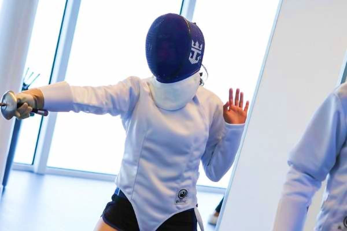 Fencer in white jacket and navy mask holding épée weapon during fencing activity on cruise ship with ocean view windows