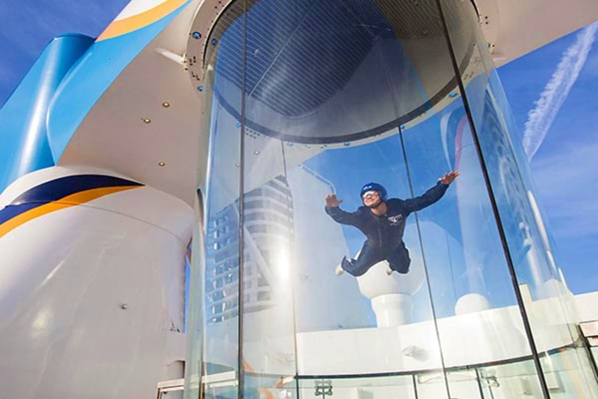 Person indoor skydiving in a transparent wind tunnel chamber on a cruise ship with city skyline visible through windows