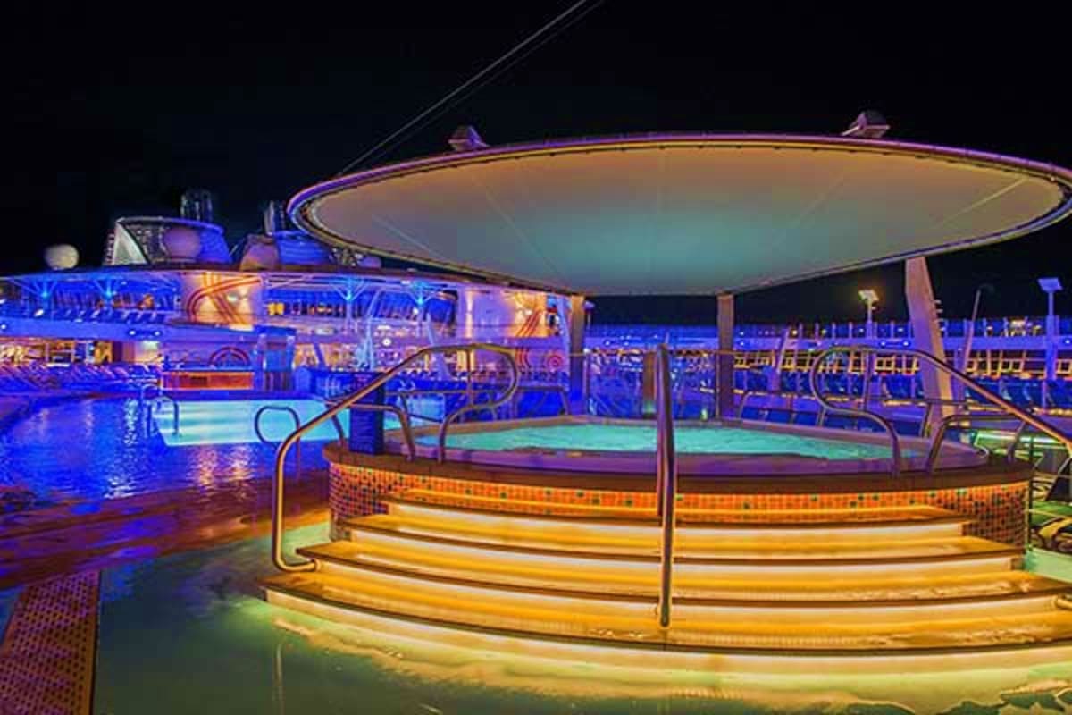 Night view of Spectrum of the Seas cruise ship pool deck with illuminated hot tubs, blue and golden lighting, and covered can