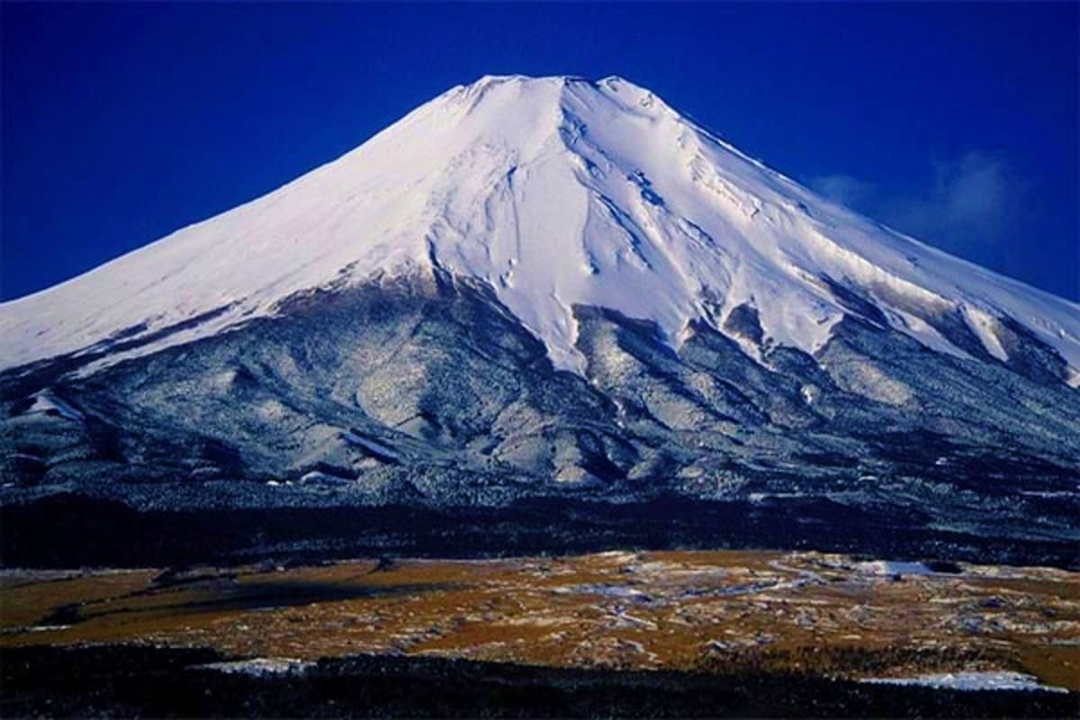 Snow-capped Mount Fuji with white peak and forested slopes against clear blue sky and brown volcanic landscape below