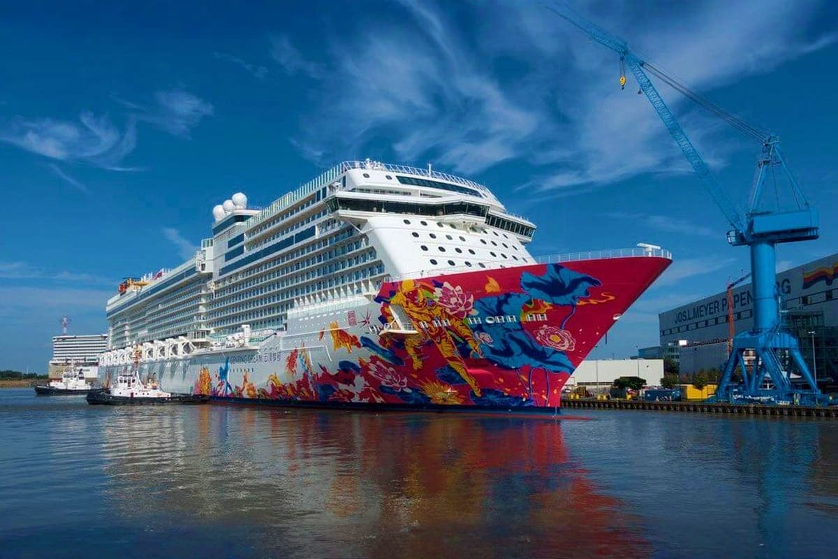 Large modern cruise ship with red and colorful artistic bow design docked at industrial port facility under blue sky