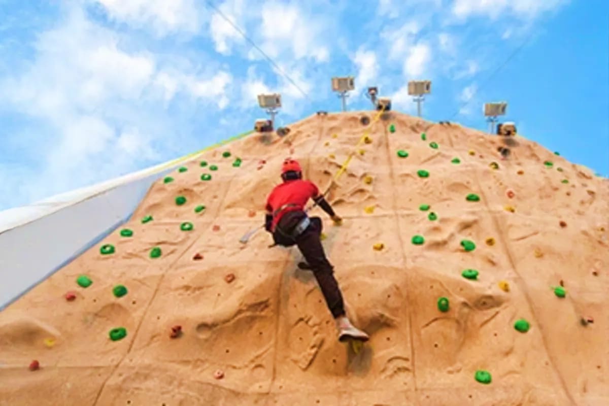 Person in red jacket rock climbing an artificial climbing wall with colorful handholds and safety lighting equipment