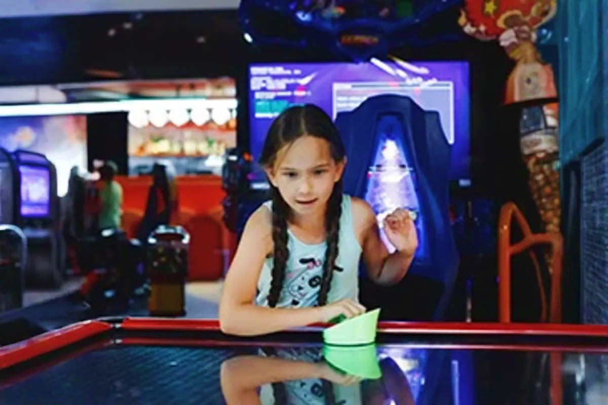Young girl playing air hockey game in cruise ship arcade with gaming machines and screens in background