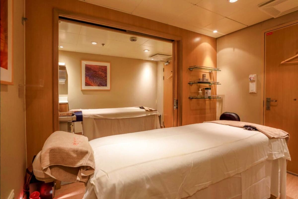 Cruise ship spa treatment room with massage bed, white linens, wooden interior paneling, and adjoining bathroom with bathtub 