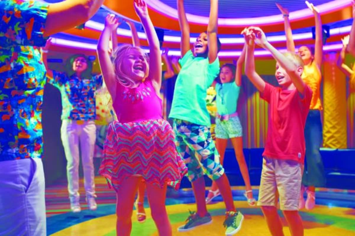 Group of children dancing and jumping with arms raised in a colorful neon-lit indoor party room with rainbow floor and bright