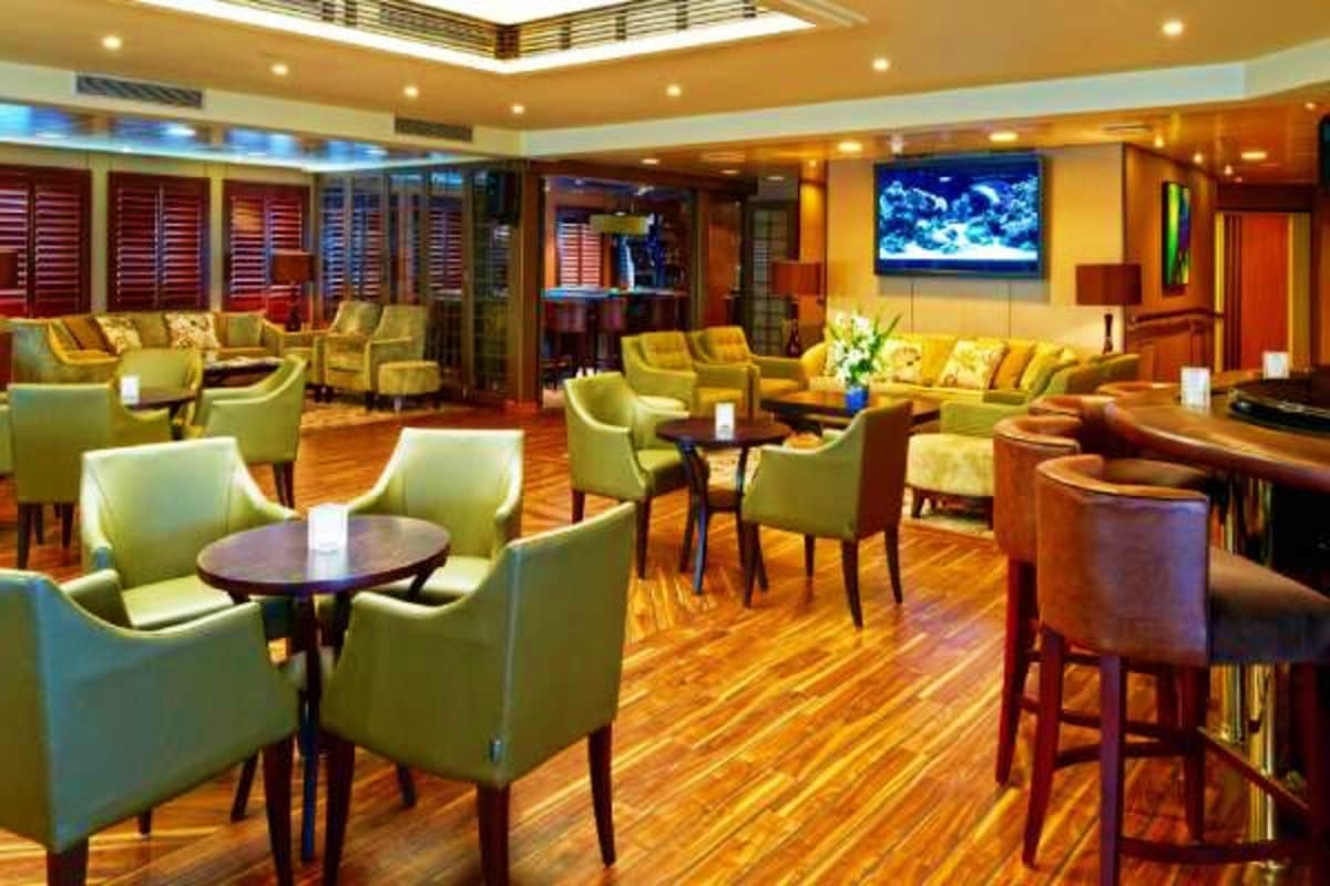 Wind Star Greek Isles Treasures Luxury cruise ship lounge with golden yellow walls, green upholstered chairs, wooden tables, aquarium display, and warm ambie
