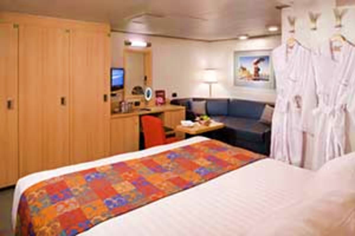 Spacious cruise ship cabin with queen bed, sitting area with black sofa, wooden wardrobe, and ocean view window with white cu