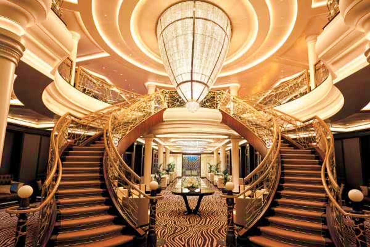 Luxury cruise ship interior with grand spiral staircase, ornate golden railings, coffered ceiling with warm lighting, and dec