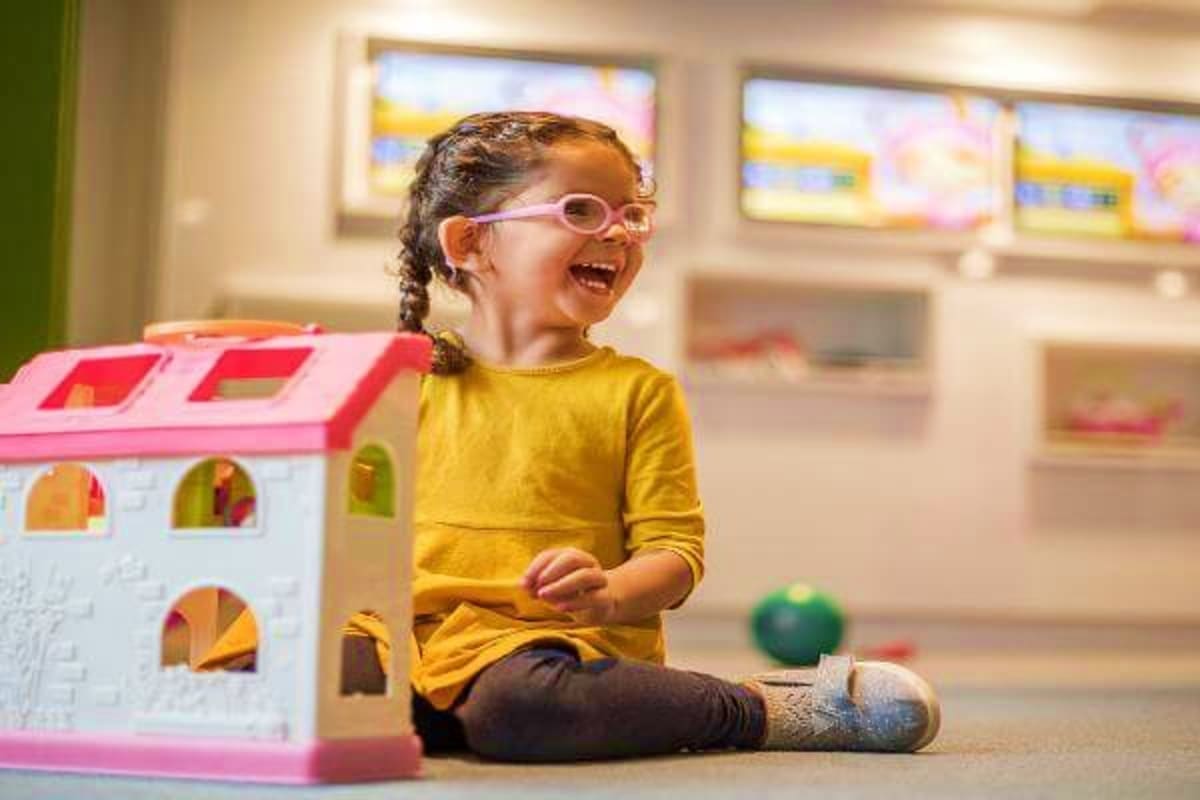 Happy young girl wearing glasses and yellow shirt playing with a pink and white toy house in an indoor playroom with educatio