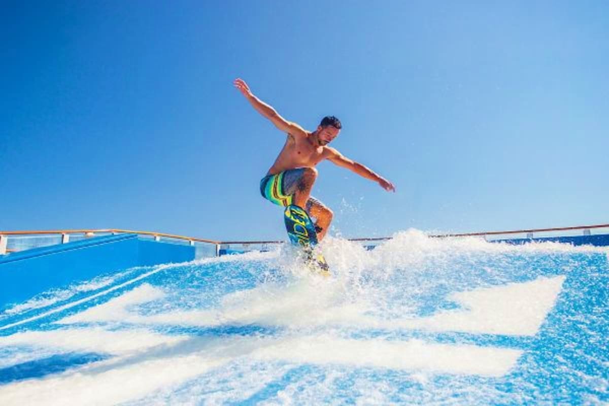 Man surfing on artificial wave at indoor surfing facility with blue walls and clear sky