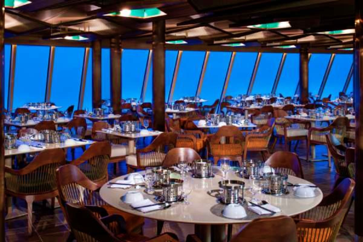 Upscale dining room on cruise ship with floor-to-ceiling windows overlooking coastal cityscape at dusk