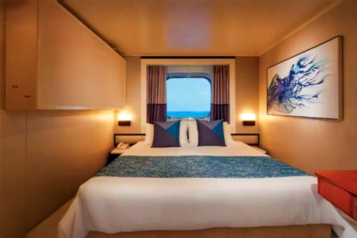 Norwegian Jade Asia Discovery Luxury cruise ship interior stateroom with ocean view window, queen bed with blue pillows, warm lighting, and modern furnishi