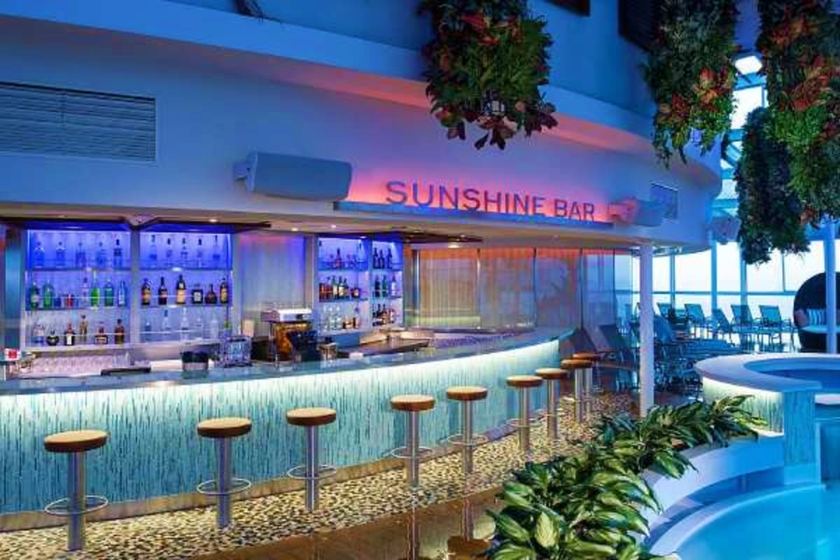 Modern Sunshine Bar on cruise ship with blue lighting, bar seating, illuminated shelving with bottles, and ocean view