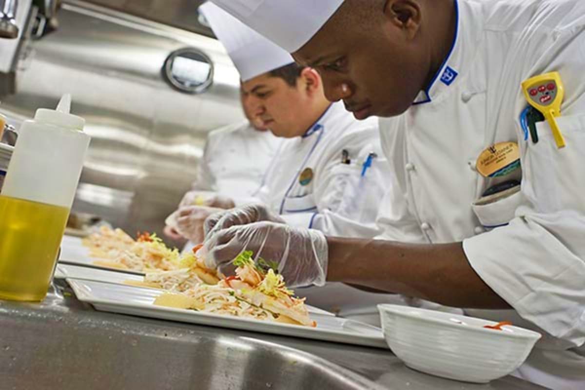 Professional chefs in white uniforms and gloves plating gourmet dishes in a modern kitchen on Anthem of the Seas cruise ship