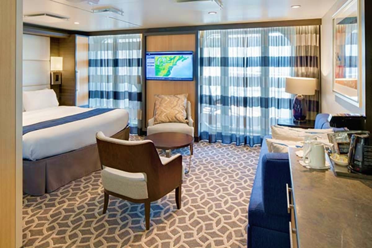 Modern cruise ship suite with queen bed, sitting area, flat-screen TV, and ocean view balcony with striped curtains