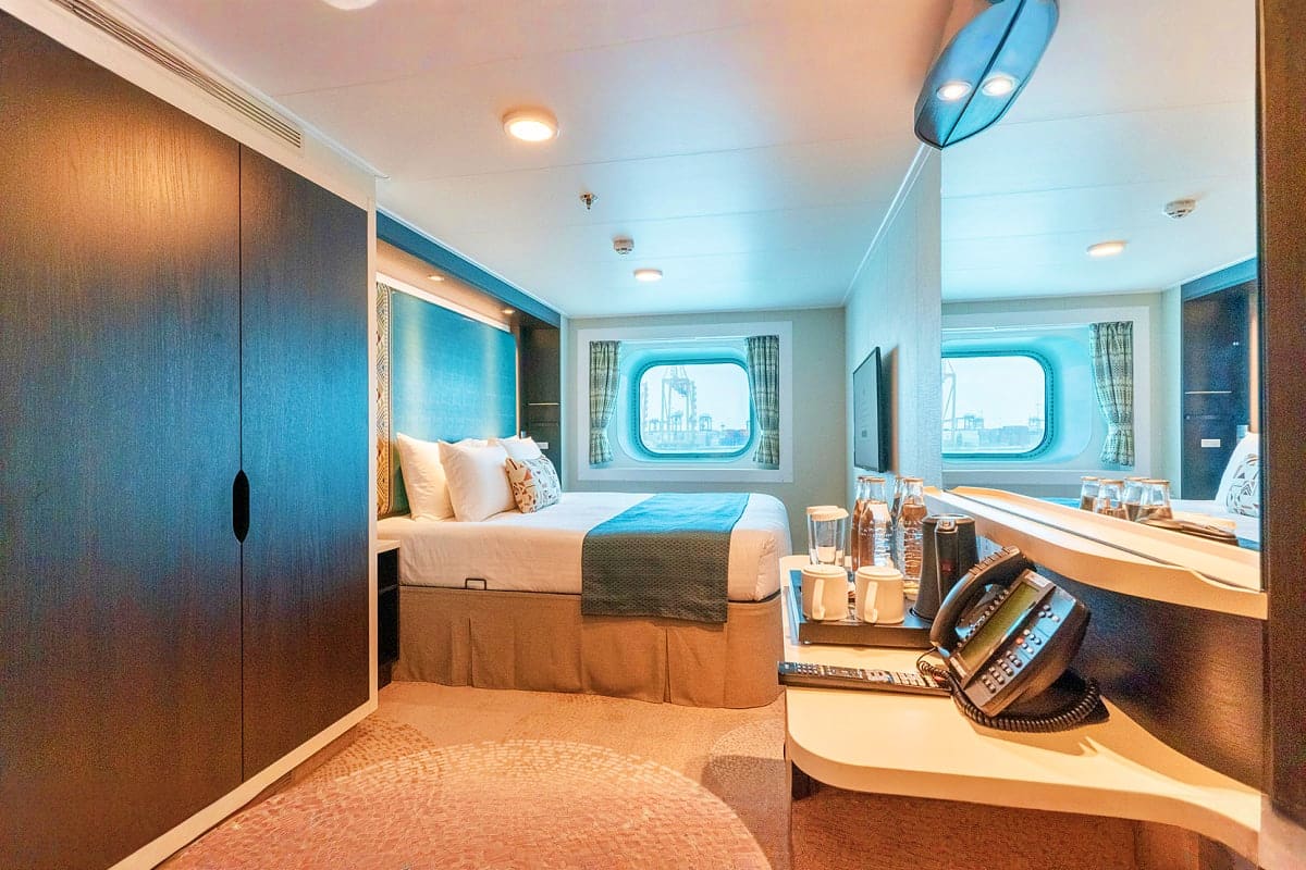 Modern cruise ship cabin with queen bed, porthole windows overlooking ocean, desk with phone, and wooden furnishings in blue 