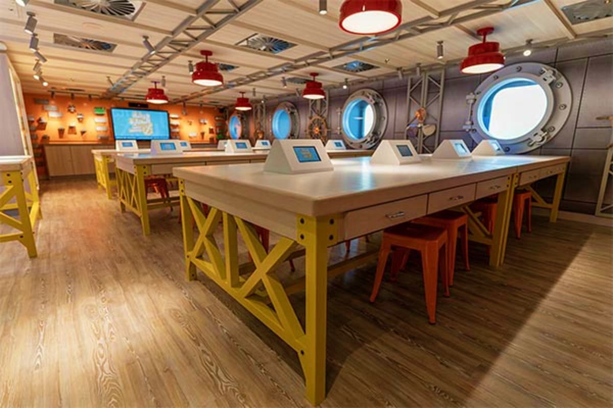 Modern cruise ship interior with white counter, yellow chairs, red pendant lights, and large circular portholes with ocean vi