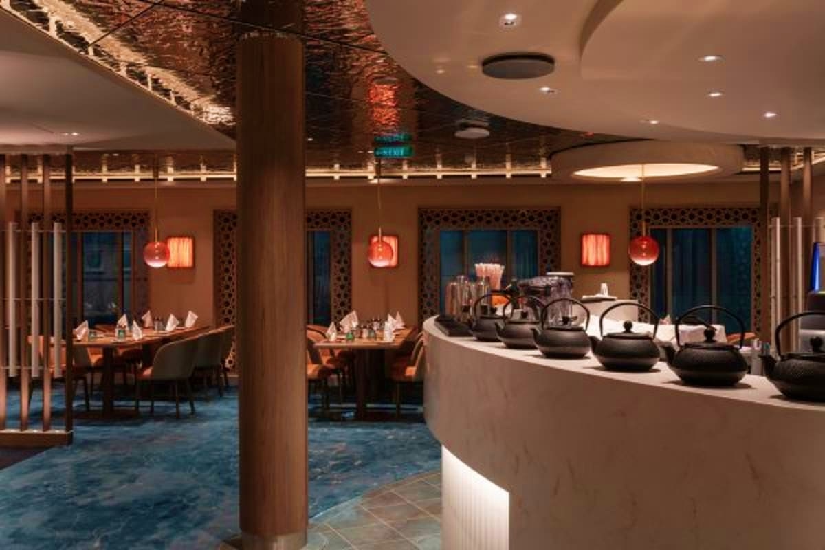 Modern cruise ship dining area with curved counter, hanging red glass lamps, wooden pillars, and elegant table seating visibl