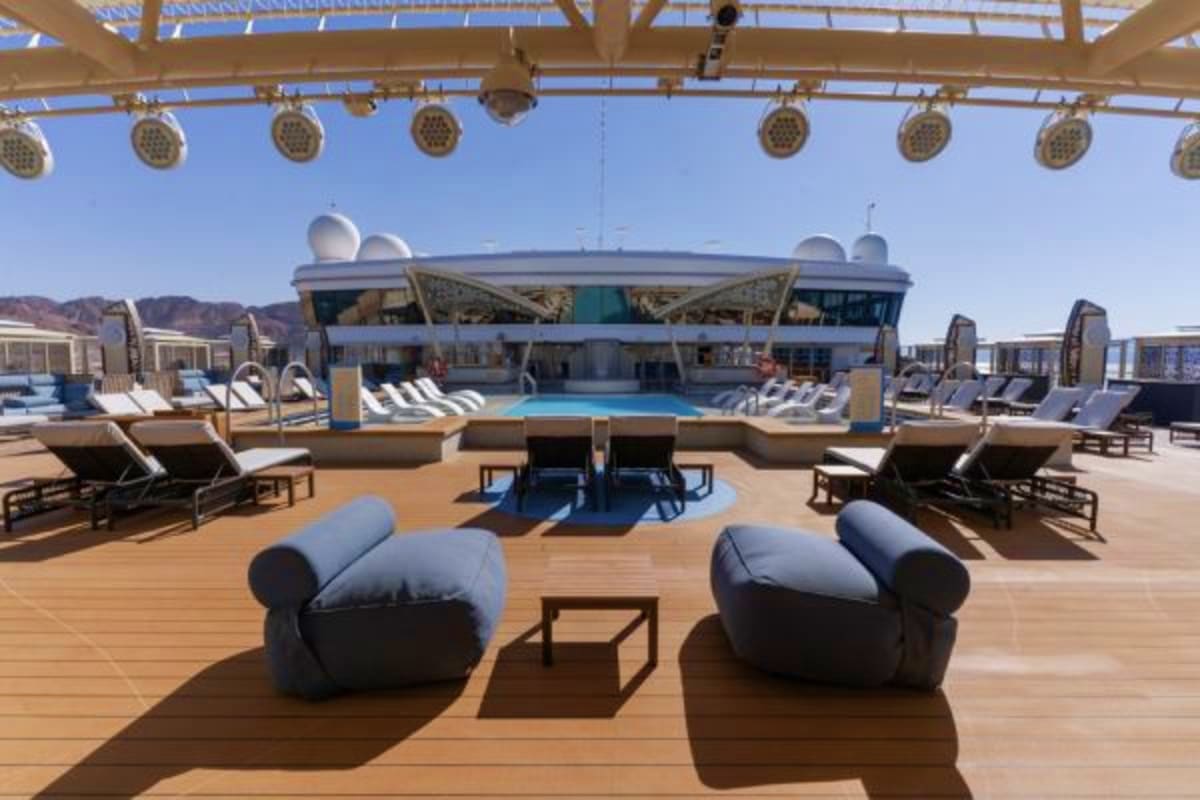 Cruise ship pool deck with blue swimming pool, lounge chairs, modern furniture, and covered dining area under clear sky