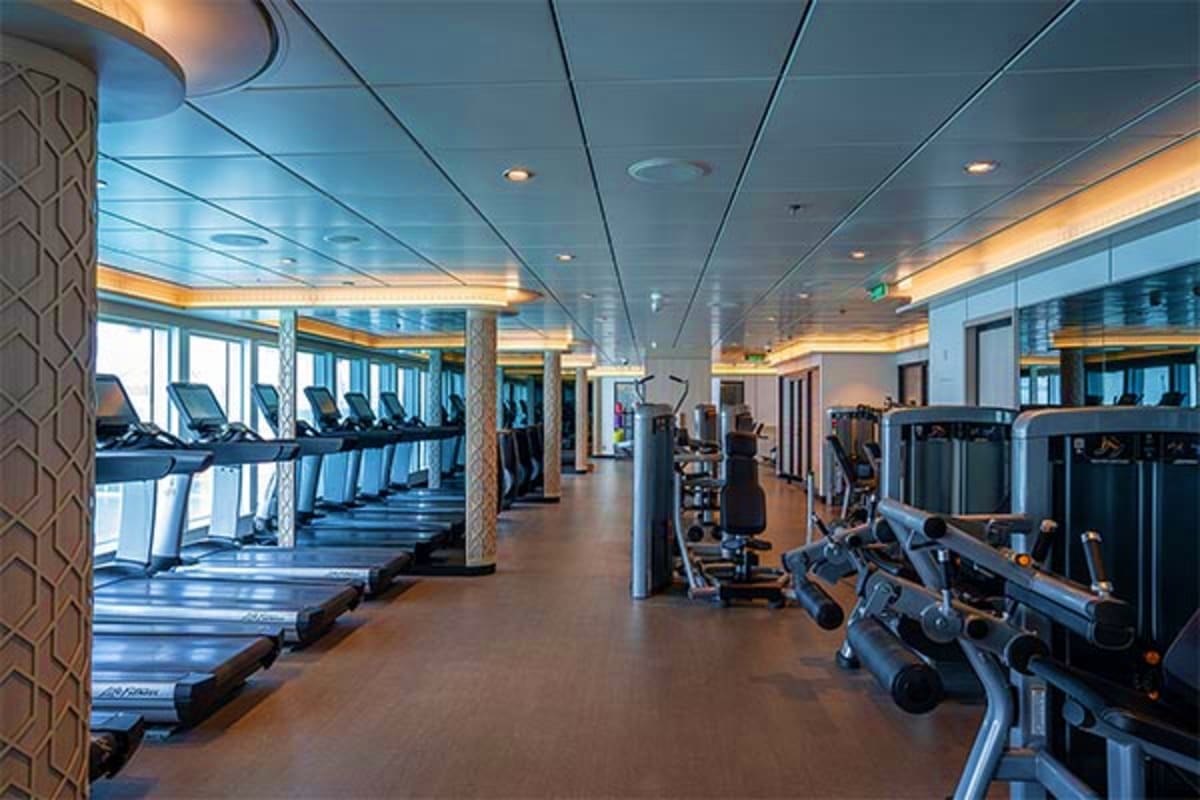 Modern ship gym with treadmills, exercise bikes, and fitness equipment overlooking ocean windows with warm ceiling lighting