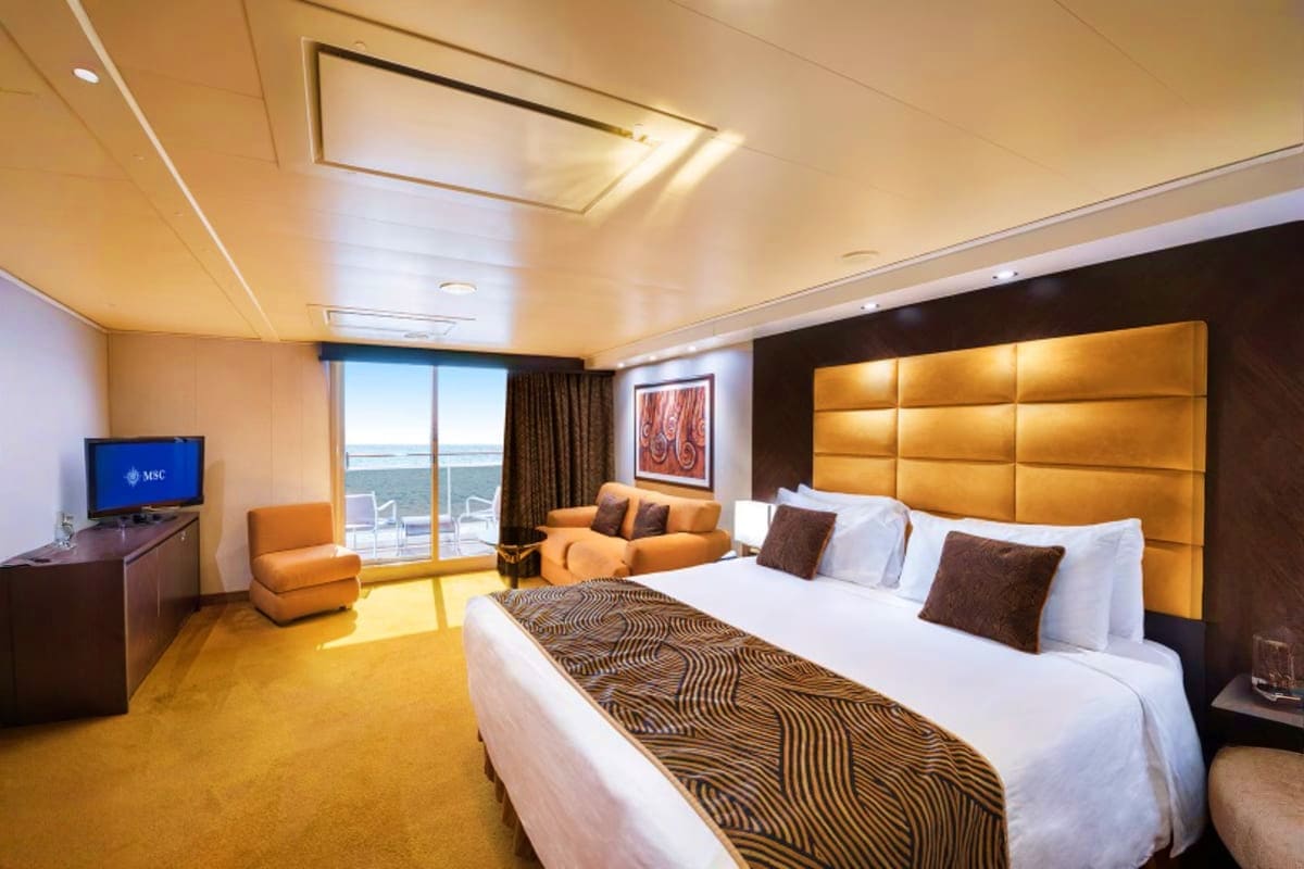 Luxury cruise ship suite with ocean view balcony, king bed with patterned runner, gold headboard, sitting area, and flat-scre