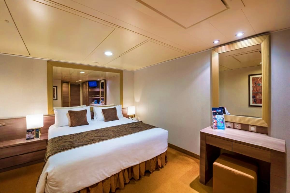 Modern cruise ship cabin with queen bed, vanity mirror, wooden furnishings, and ensuite bathroom with shower
