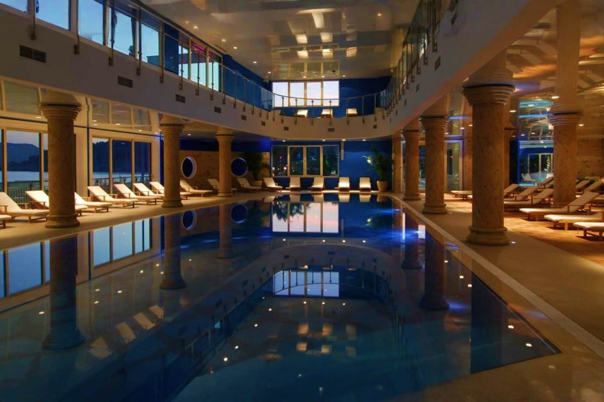 Luxurious indoor swimming pool with blue water, wooden columns, lounge chairs, and large windows overlooking the sea on MSC S