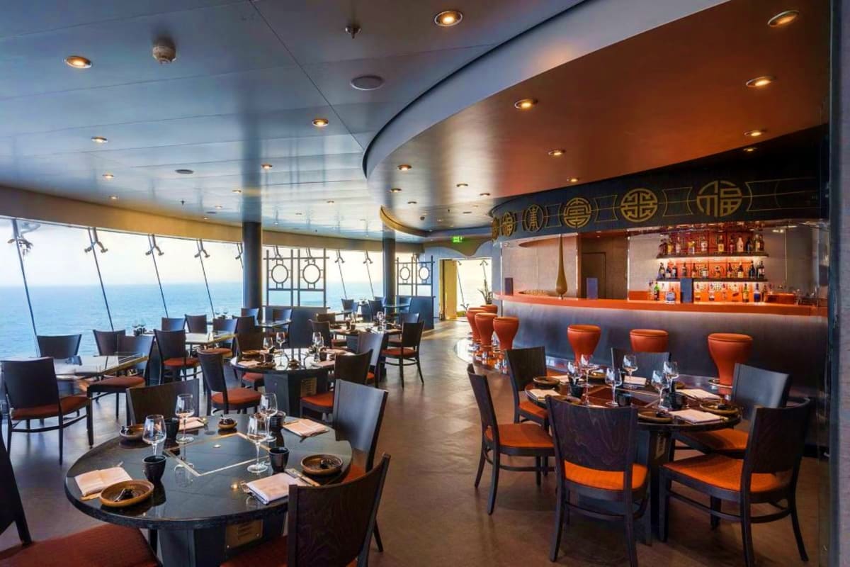 Modern cruise ship dining room with round tables, orange chairs, floor-to-ceiling ocean views, curved copper ceiling, and bar