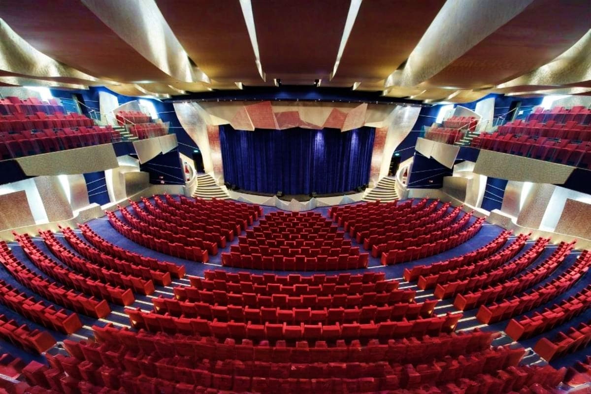 Modern theater interior with rows of red seats facing a stage with blue curtains and dramatic ceiling lighting