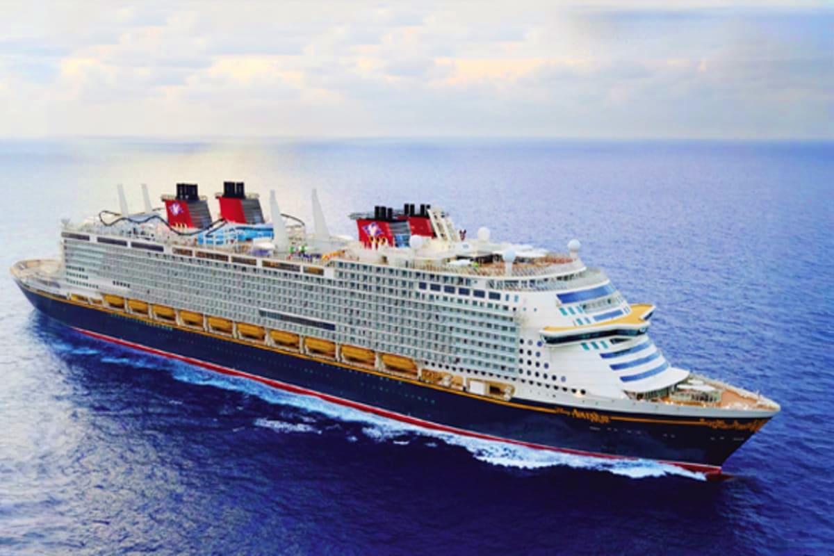 Three Nights Magical Disney Getaway Large white Disney cruise ship with red funnels sailing on blue ocean water under partly cloudy sky