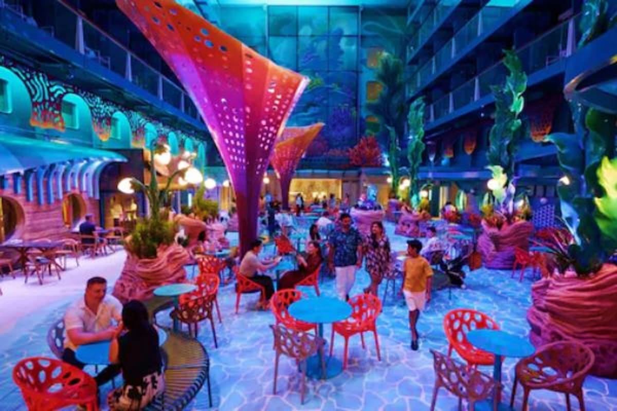 Vibrant indoor atrium with neon lighting, red and blue furniture, tropical plants, and guests dining and walking in a modern 