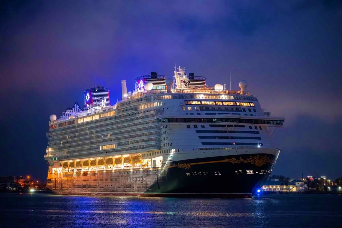 Large illuminated cruise ship docked at night with colorful lights reflecting on dark water, multiple decks visible against t