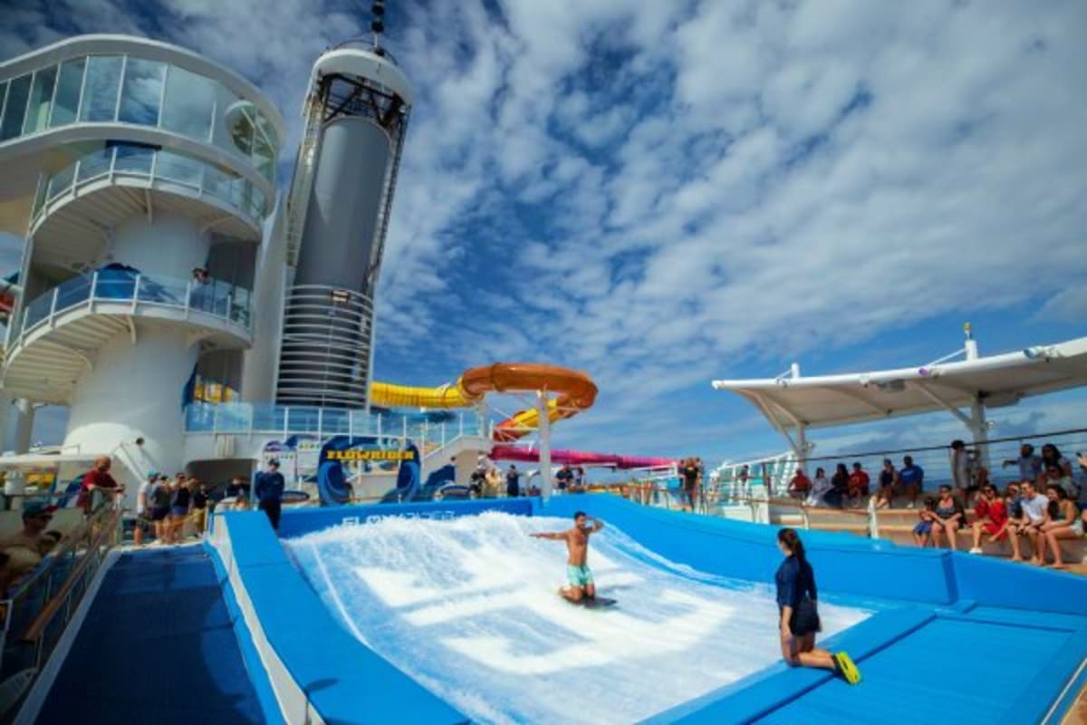 Passengers enjoying waterslides and wave pool on cruise ship deck with funnel and observation tower against cloudy sky