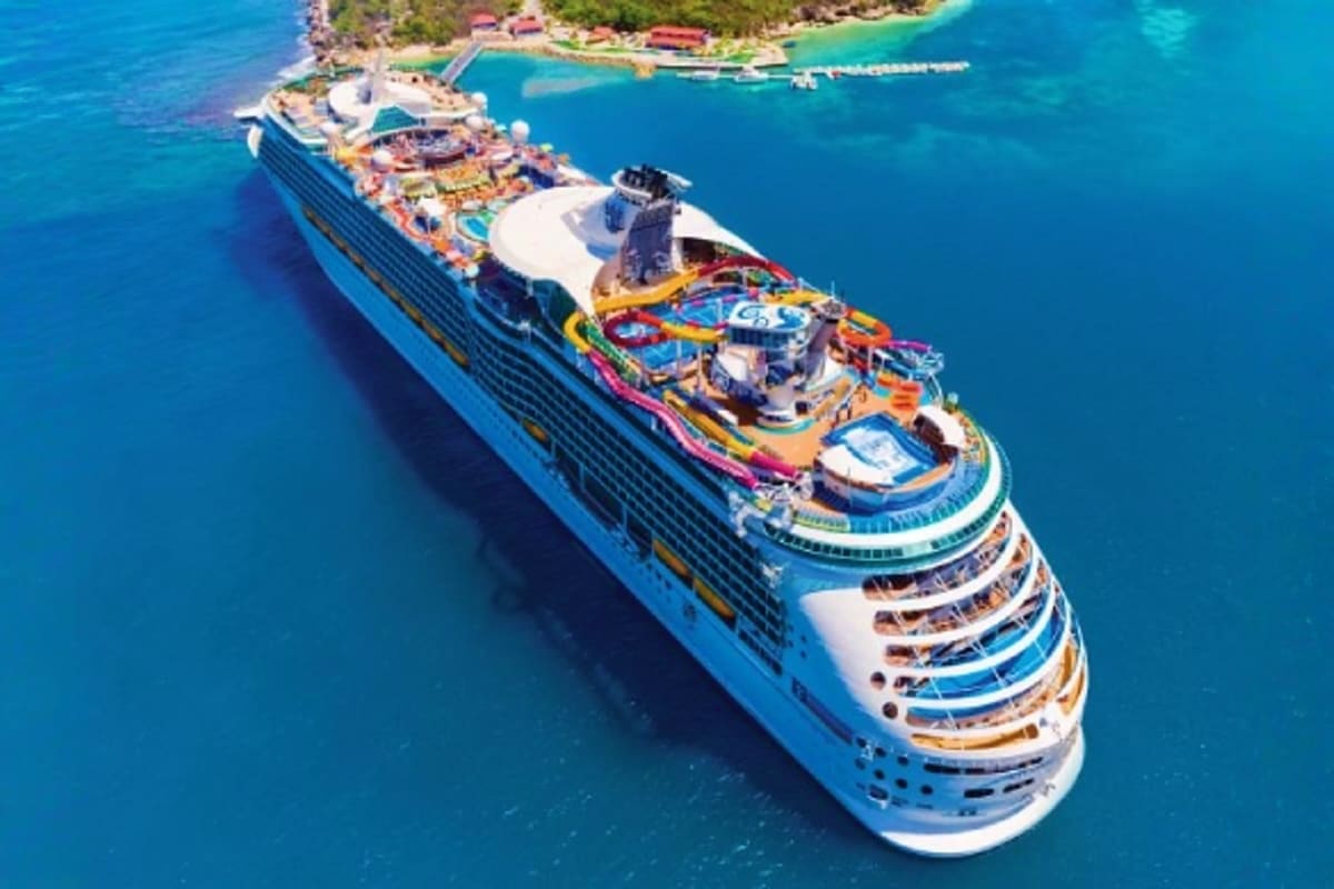 Large blue and white cruise ship with multiple decks and colorful amenities anchored in turquoise tropical waters near a coas