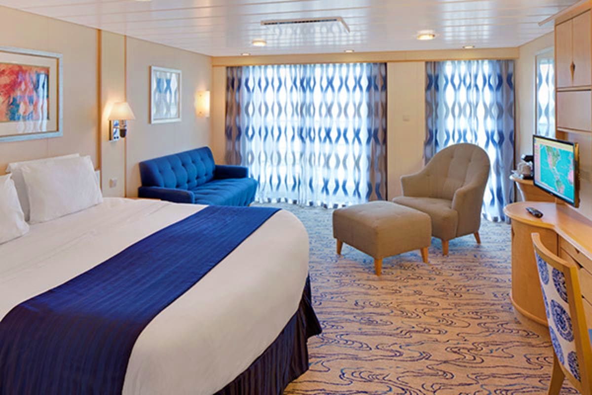 Luxury cruise ship cabin with queen bed, blue sofa, beige armchair, patterned curtains, and ocean view balcony