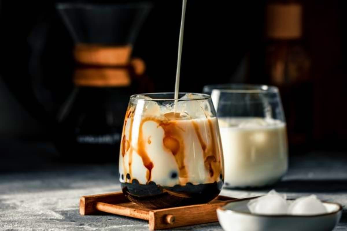 Iced coffee drink with milk being poured into glass, caramel drizzle, wooden coaster, milk pitcher and coffee equipment in ba