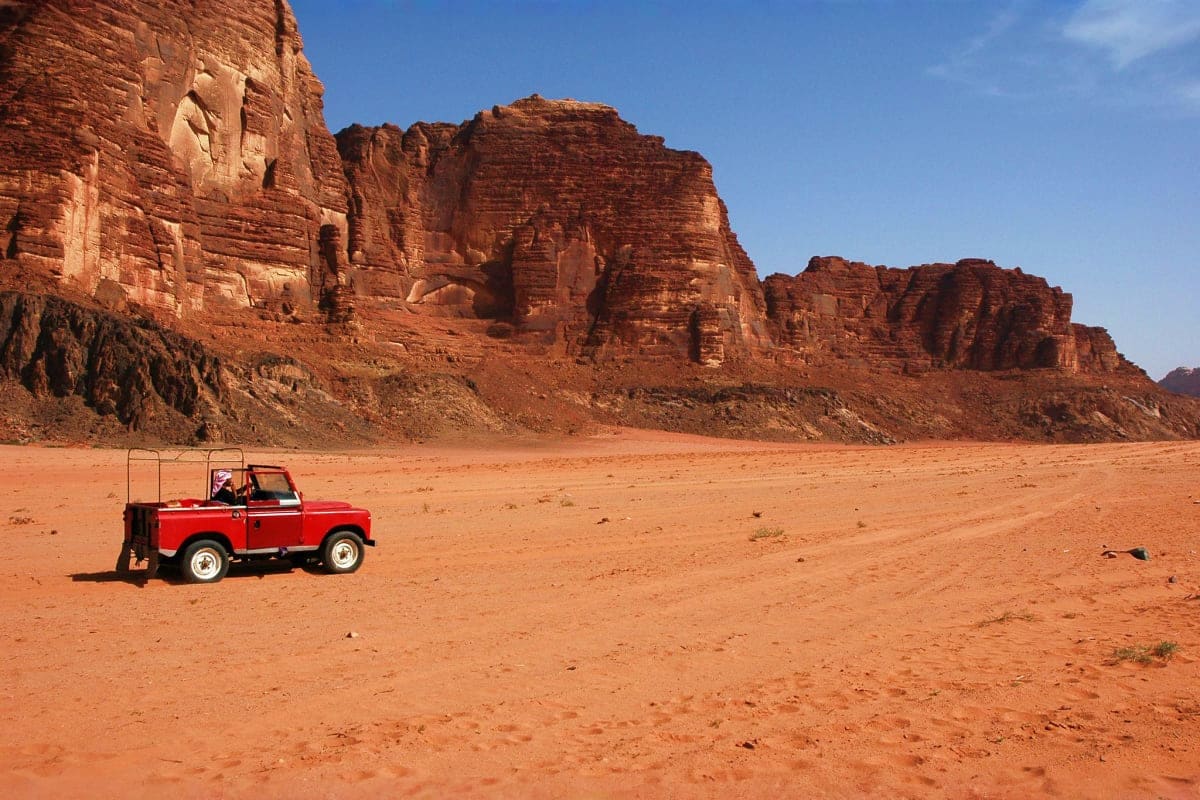 Red Land Rover parked in Wadi Rum desert with towering red sandstone cliffs under blue sky