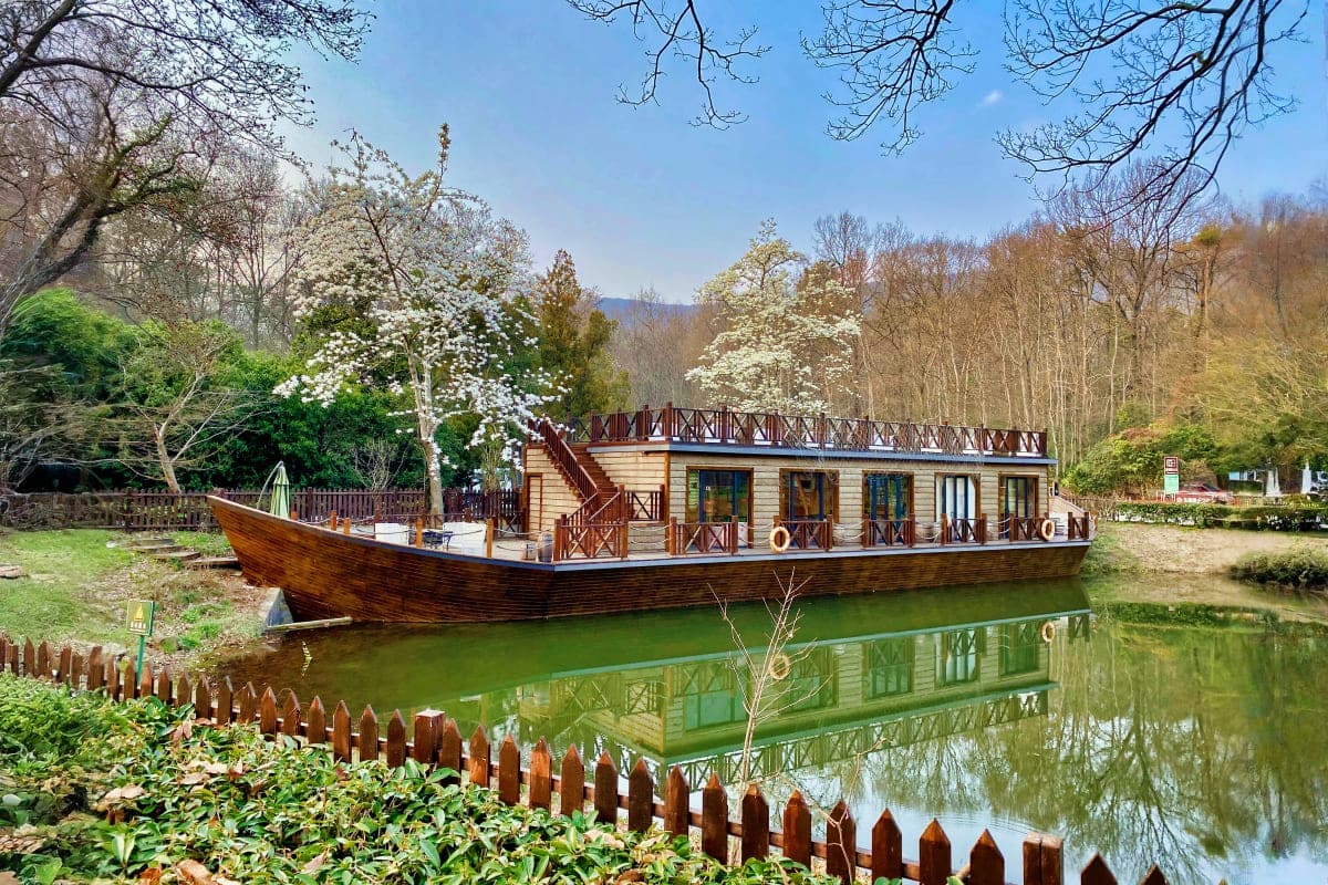 Wooden boat-shaped building on a canal surrounded by blooming spring trees and wooden fence, reflecting in calm green water