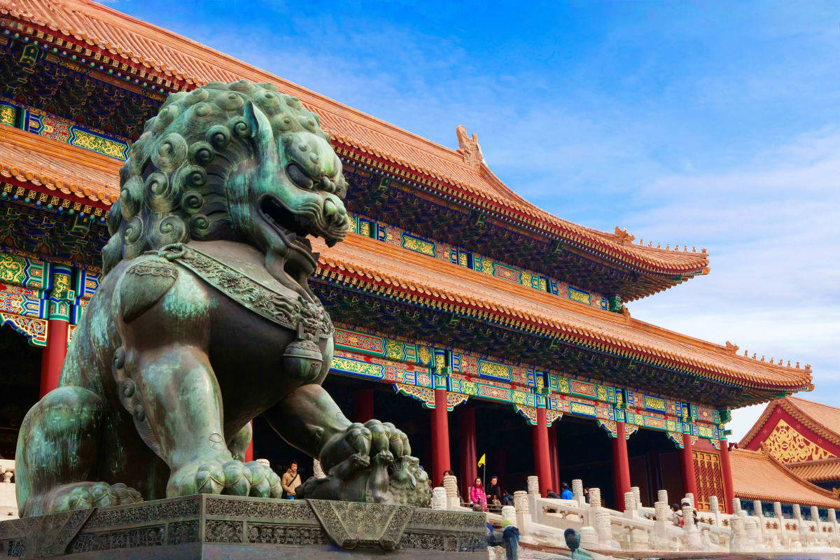 Bronze lion statue guarding the entrance to a traditional Chinese temple with ornate golden-glazed tile roofs and red pillars