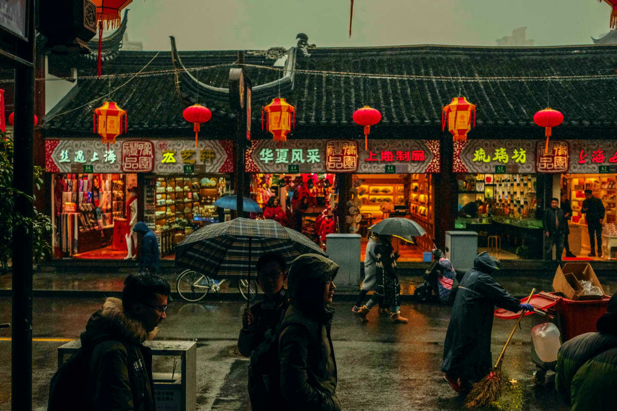 Rainy street scene in traditional Chinese town with red lanterns, shop fronts with Chinese characters, and pedestrians holdin