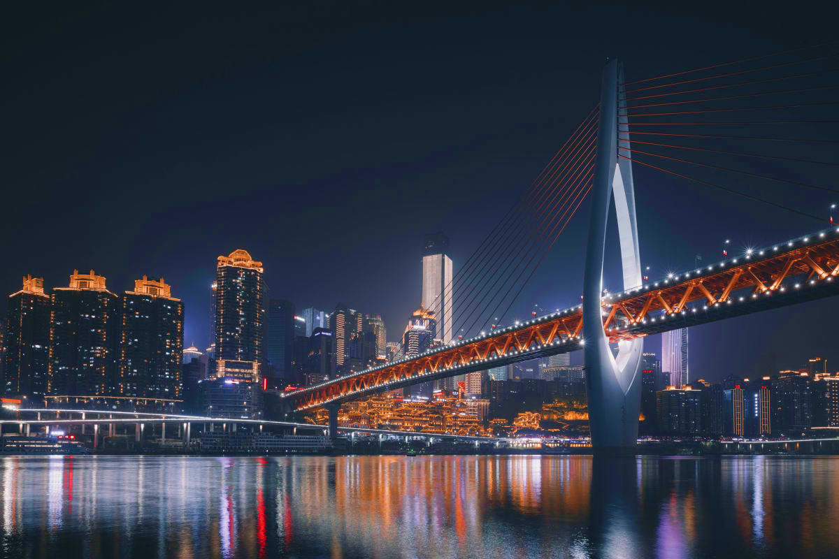 Modern illuminated bridge spanning river at night with city skyline and lit skyscrapers reflected in water
