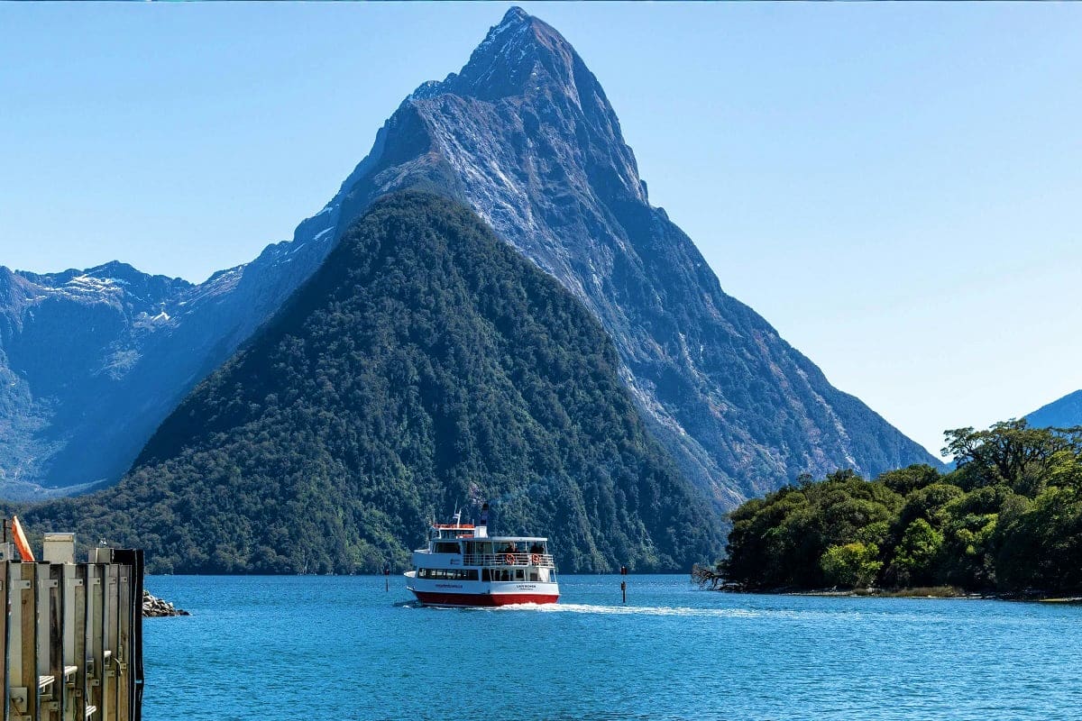South Island Tour of New Zealand – 5 nights & 6 days Tourist cruise boat on turquoise water with dramatic forested mountain peak in Milford Sound, New Zealand
