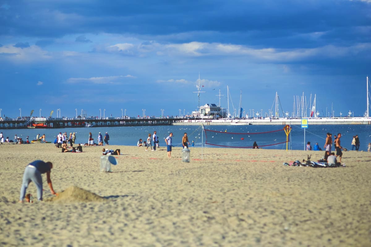 Busy sandy beach with swimmers and beachgoers, sailboats in harbor, and pier with buildings under blue sky with clouds