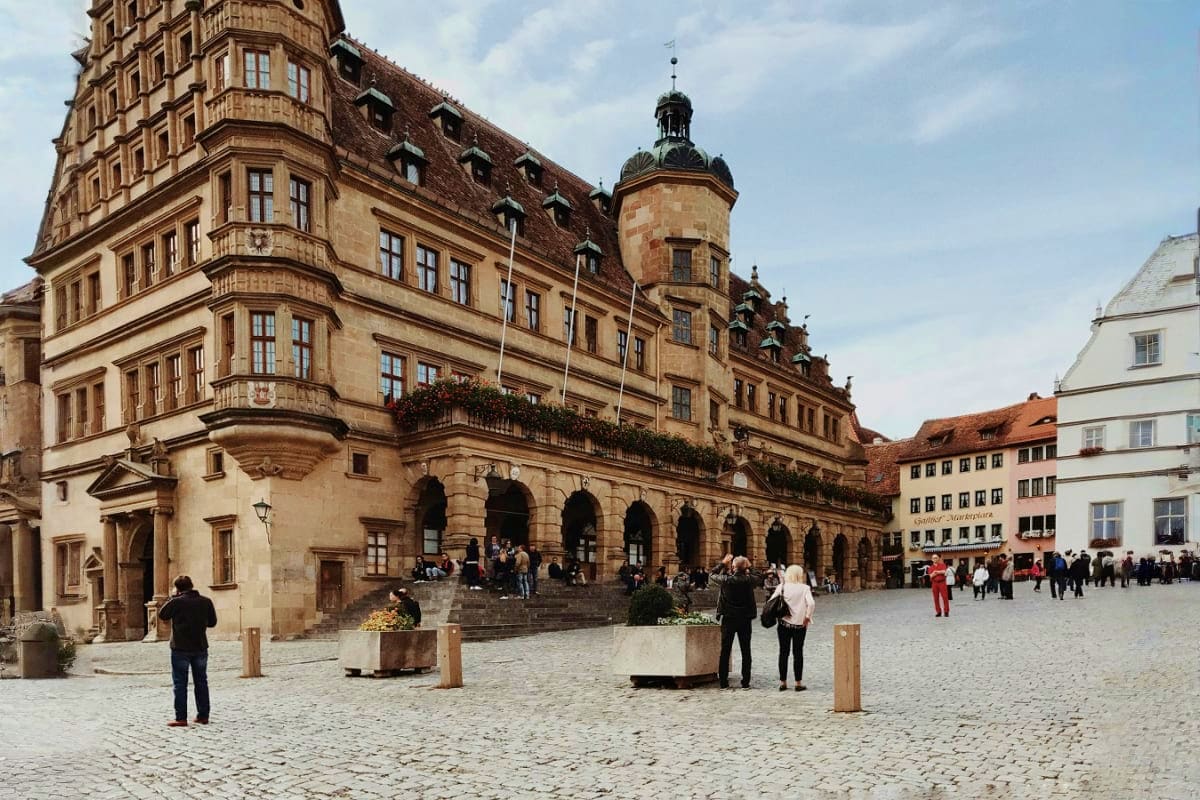 Historic European town square with Renaissance architecture, ornate sandstone building with tower, and visitors exploring cob