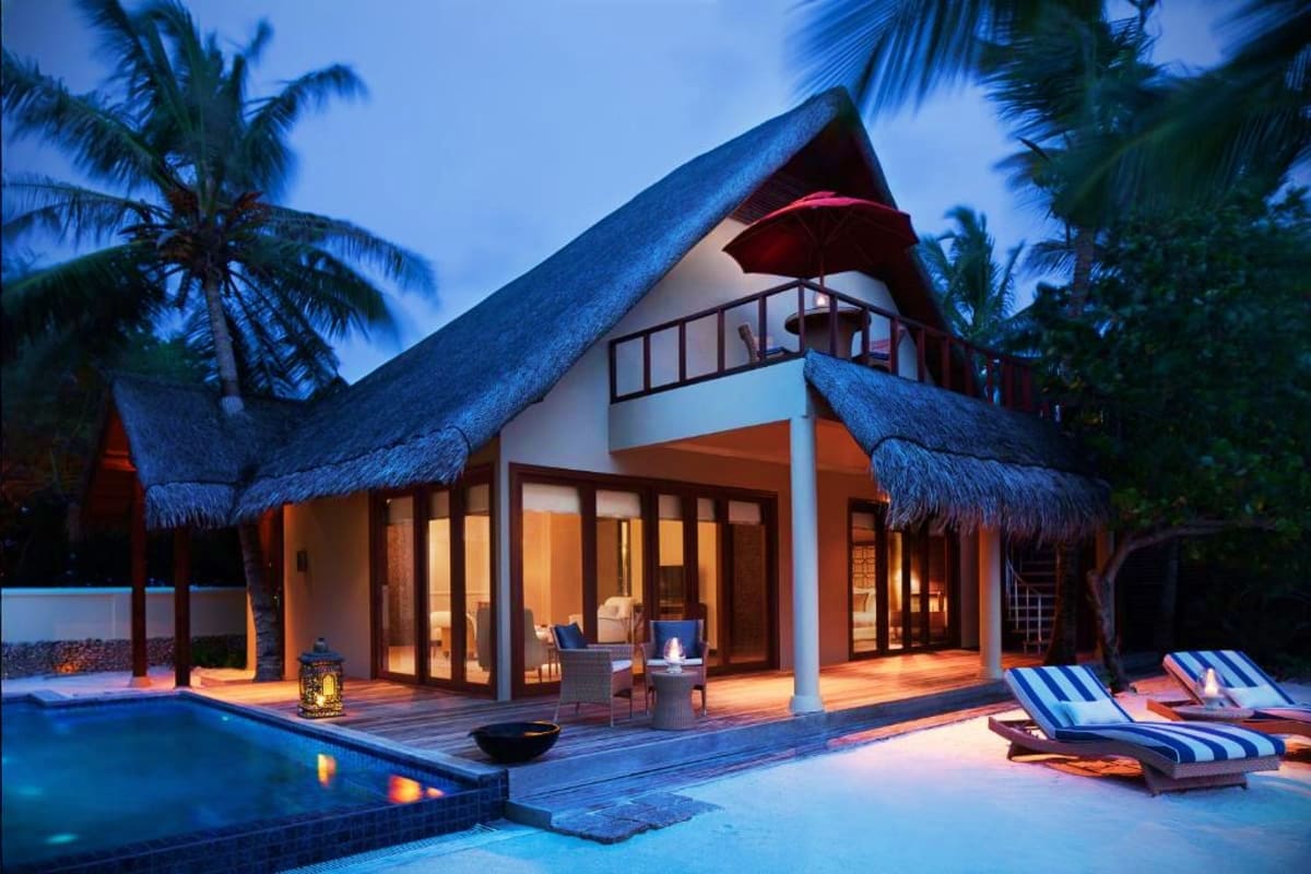 Luxury tropical villa with thatched roof, illuminated interior, private pool, lounge chairs, and palm trees at dusk