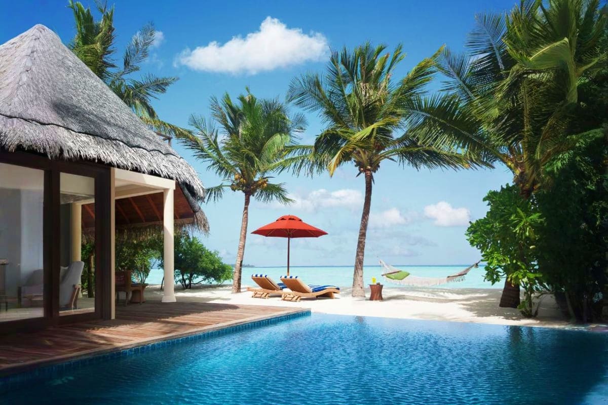 Luxury beachfront villa with thatched roof, infinity pool, palm trees, and turquoise ocean view with lounge chairs and red um