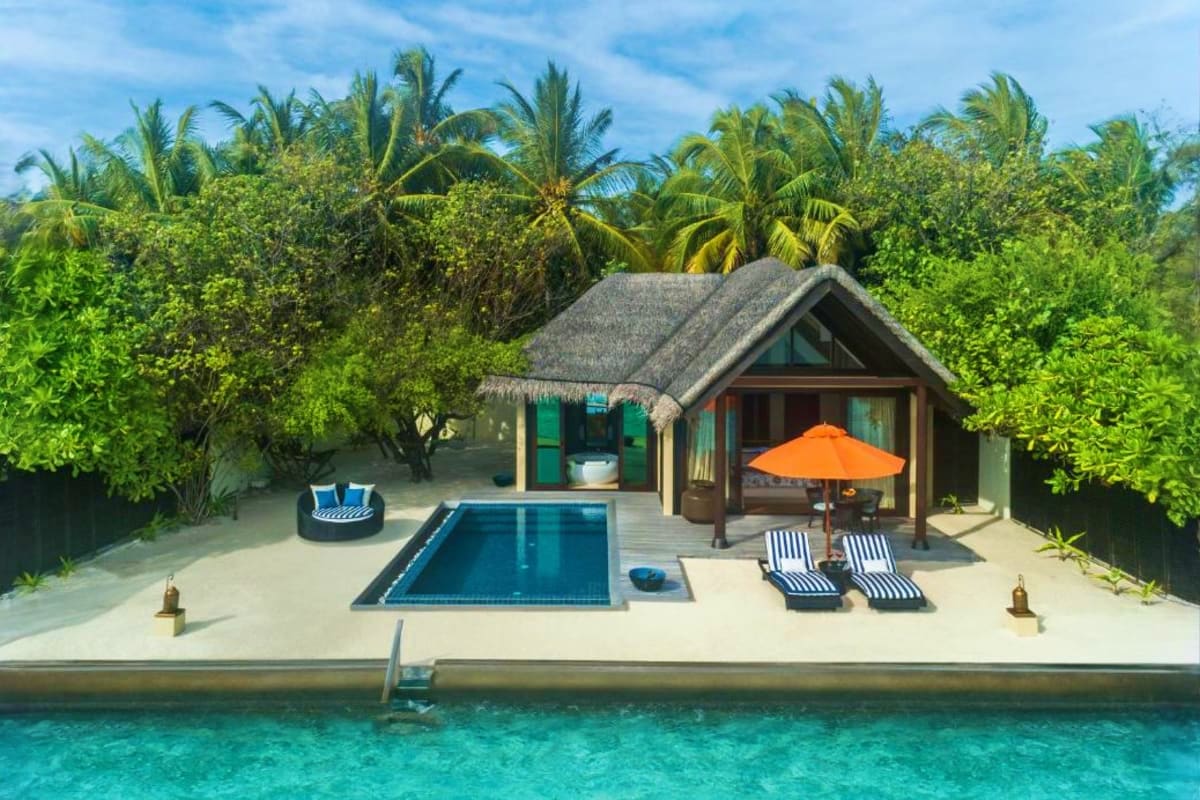 Luxury beach villa with private pool, thatched-roof bungalow, lounge chairs with orange umbrella, and tropical palm trees ove