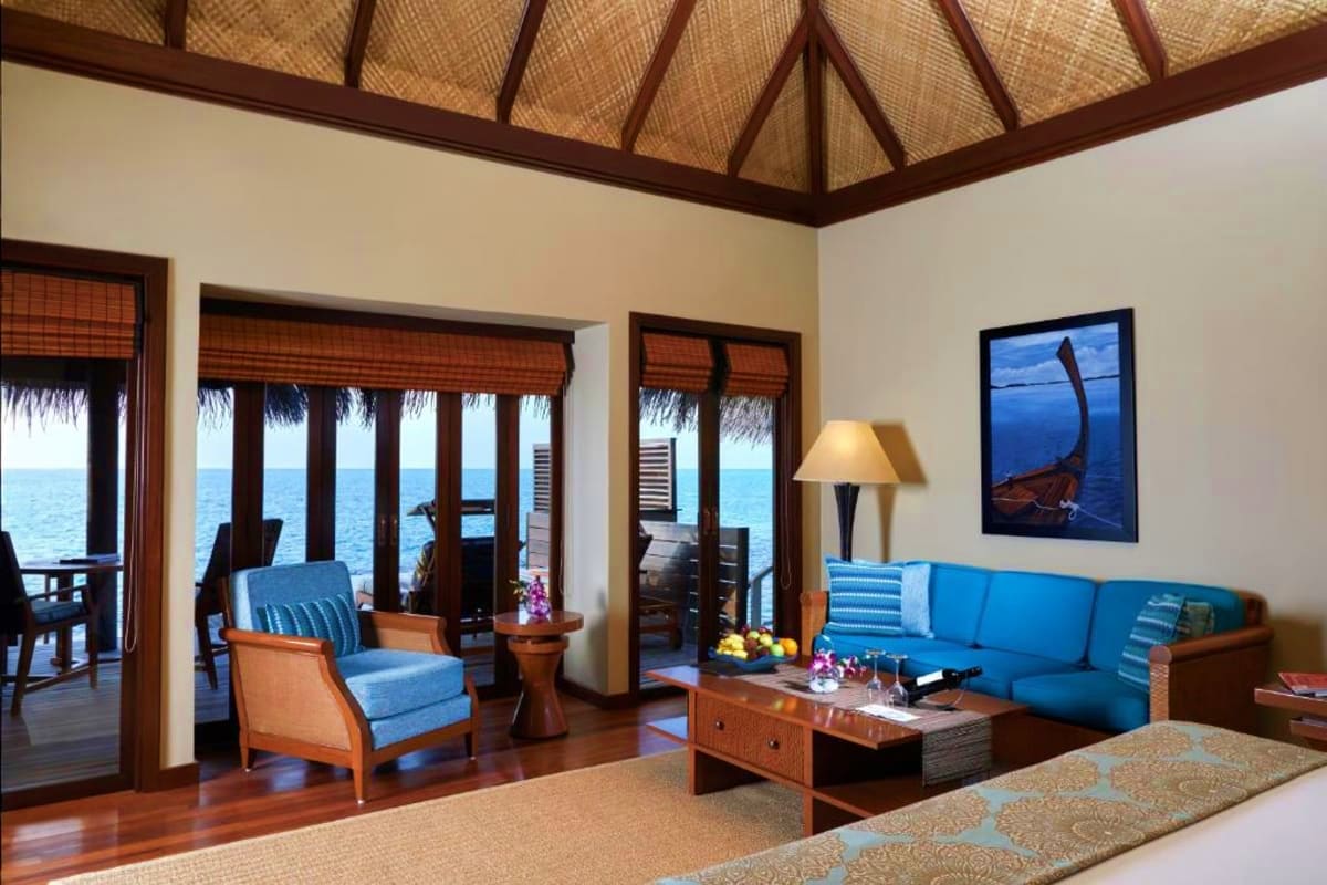Luxury overwater bungalow interior with ocean views, wooden furniture, blue upholstered sofa, and thatched ceiling at tropica
